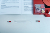 Ein offenes Set mit einem roten Koffer enthält diverses Zubehör sowie eine Anleitung zu physikalischen Experimenten. Im Vordergrund liegen zwei kleine Magnete und ein leeres Reagenzglas auf einer weißen Fläche.
